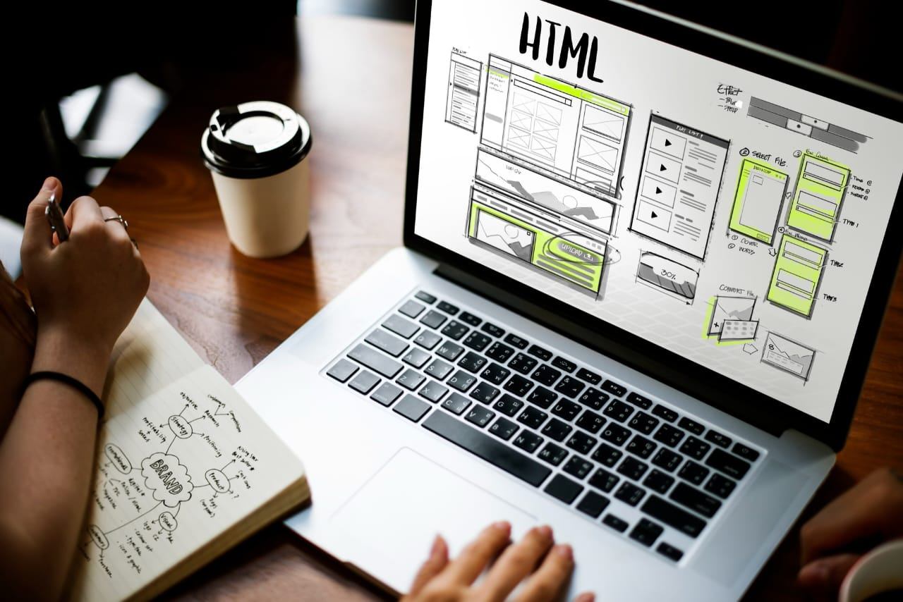 Laptop mostrando el boceto de una página web junto a un cuaderno de ideas
